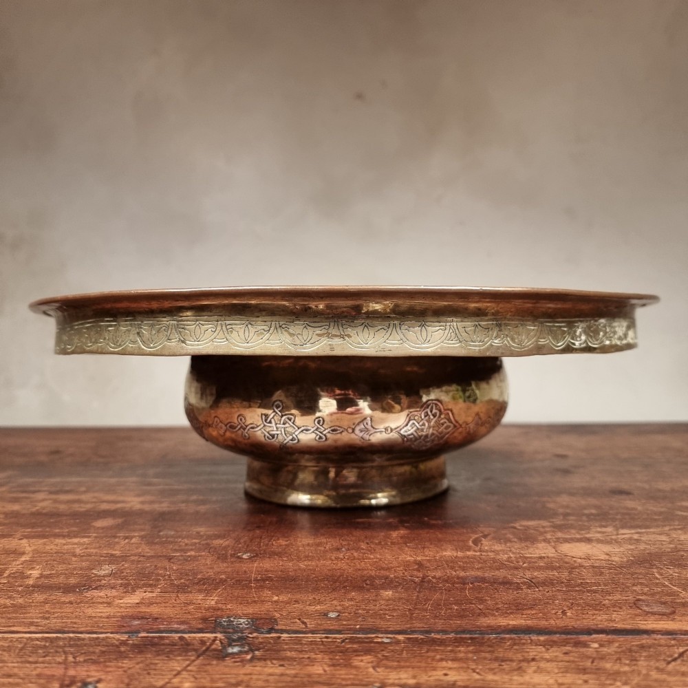 A circular 19th-century Islamic brass wash bowl featuring intricate Arabic calligraphic script, geometric arabesque patterns, pierced metalwork, and silver and copper inlay. View V