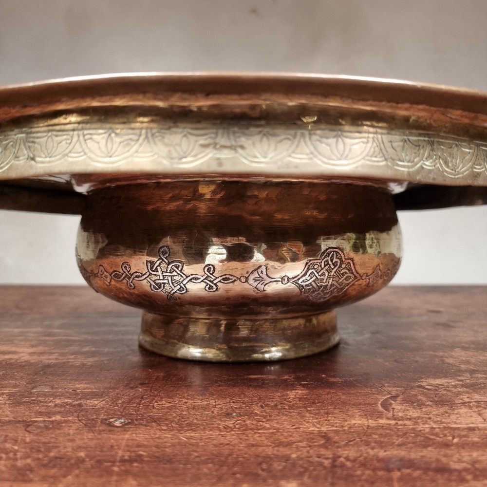 A circular 19th-century Islamic brass wash bowl featuring intricate Arabic calligraphic script, geometric arabesque patterns, pierced metalwork, and silver and copper inlay. View VI