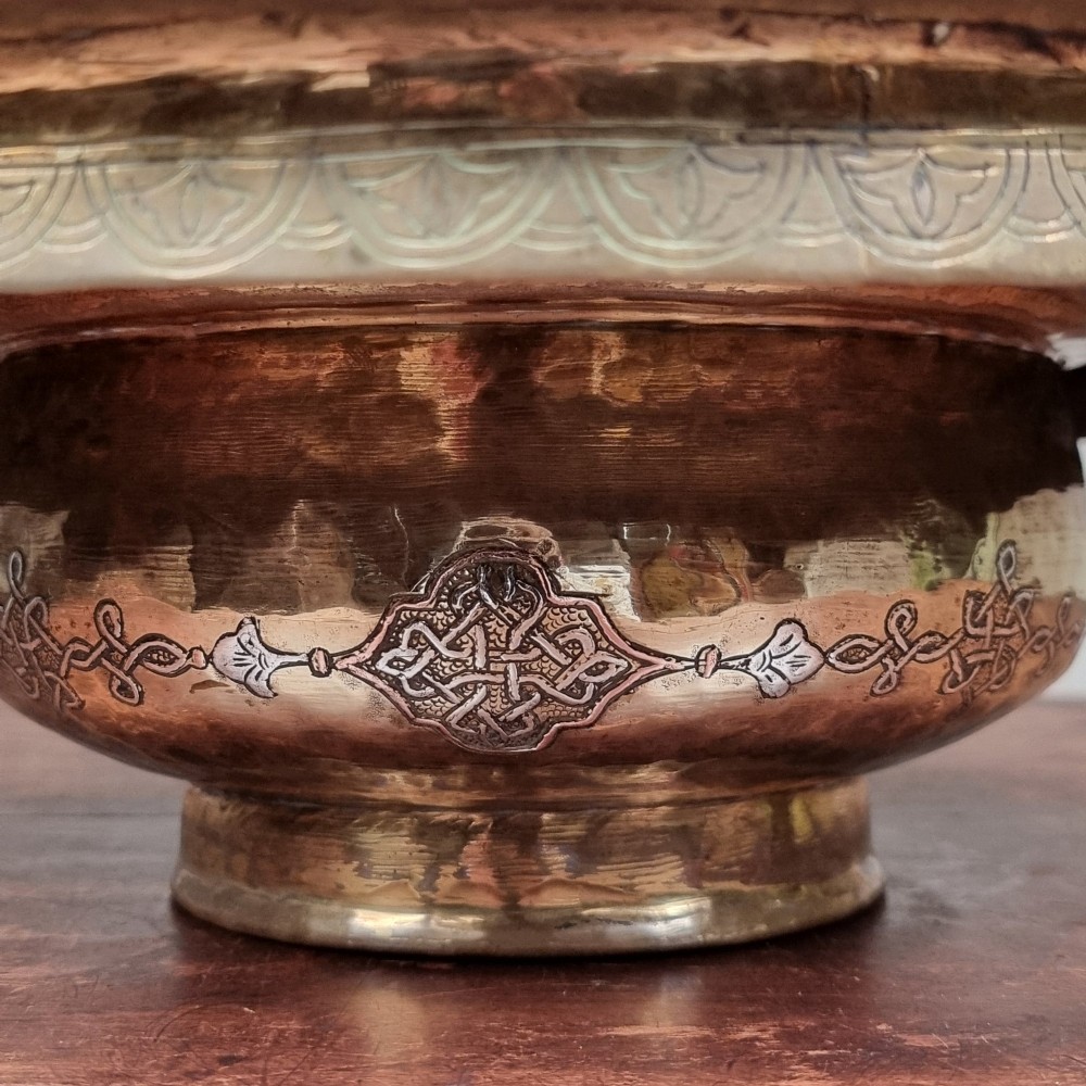 A circular 19th-century Islamic brass wash bowl featuring intricate Arabic calligraphic script, geometric arabesque patterns, pierced metalwork, and silver and copper inlay. View VIII