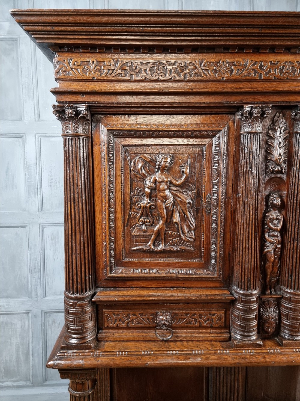 A dark carved oak cupboard featuring two relief-carved doors with figure motifs, upper drawers with ring handles, fluted columns, and an open lower tier. View II