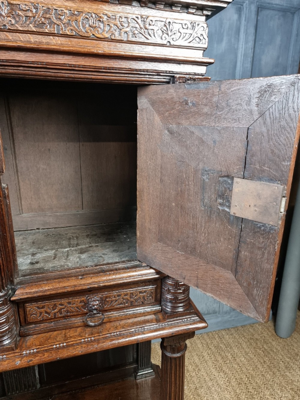 A dark carved oak cupboard featuring two relief-carved doors with figure motifs, upper drawers with ring handles, fluted columns, and an open lower tier. View V