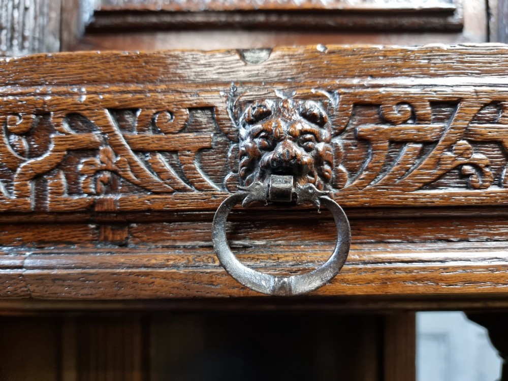 A dark carved oak cupboard featuring two relief-carved doors with figure motifs, upper drawers with ring handles, fluted columns, and an open lower tier. View VII
