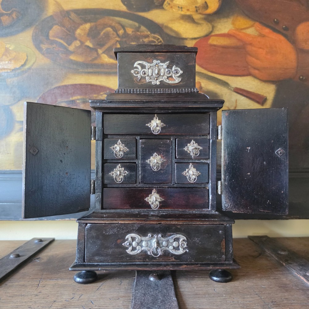 A tiered 18th-century walnut jewellery cabinet featuring ornate silvered metal hinges, a central latch mechanism, a lower drawer, and turned bun feet. View VI