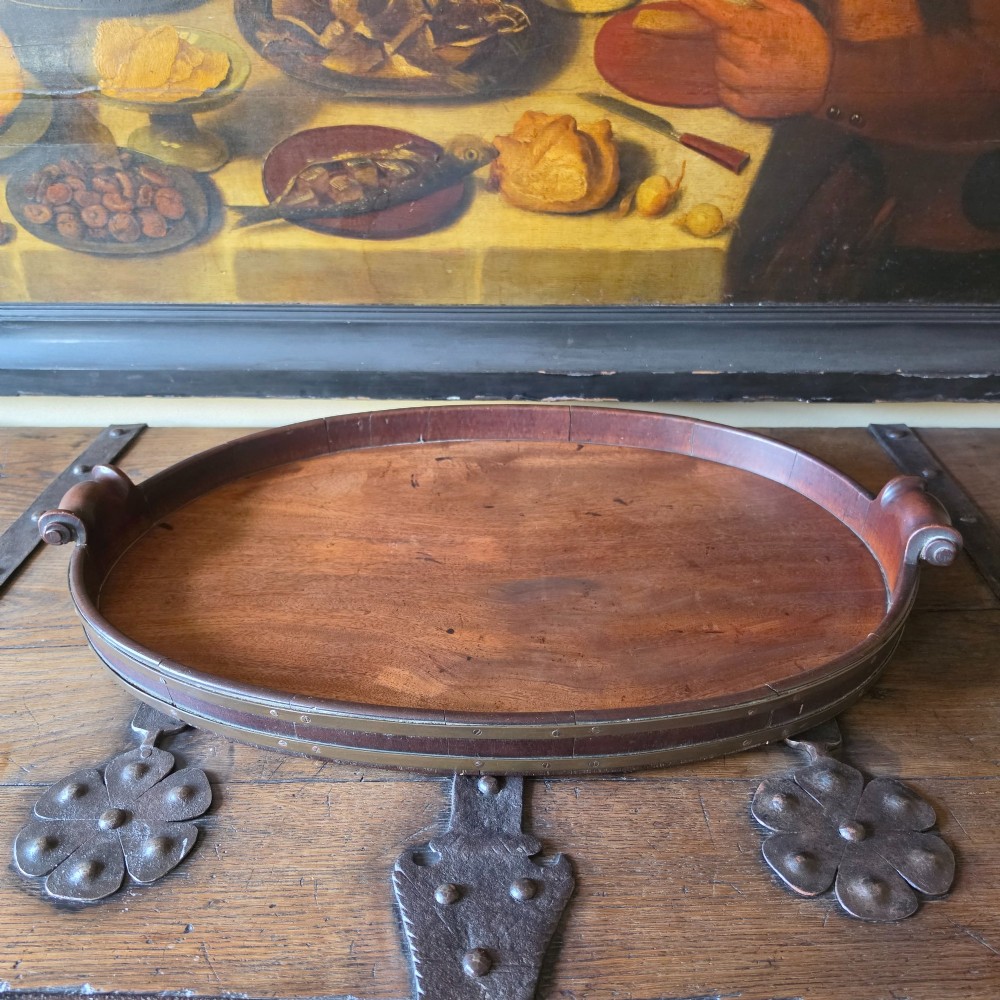 An 18th-century oval mahogany serving tray featuring a raised gallery edge with brass rim binding and carved scroll handles.