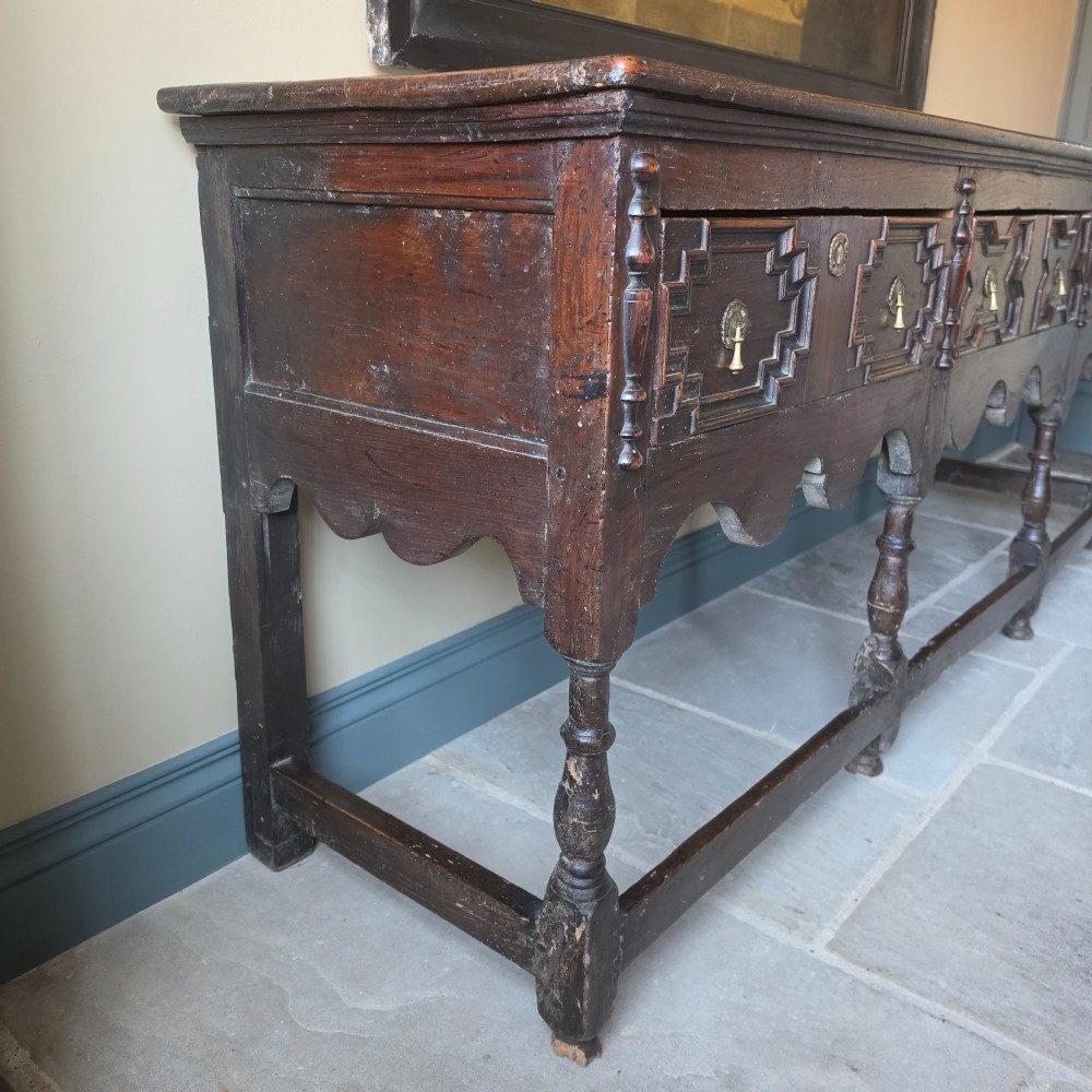 A long 17th-century dark oak dresser base featuring geometric moulded drawers, turned legs, and a shaped apron. View XII