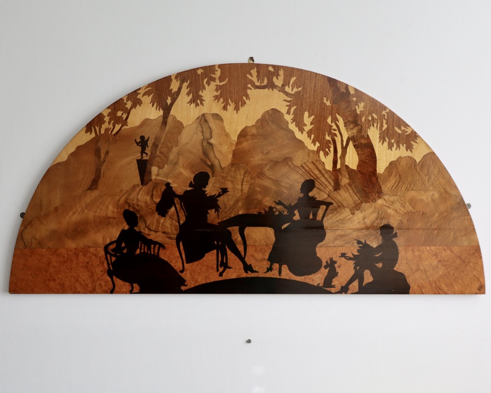A semi-circular wooden panel featuring intricate marquetry depicting silhouettes of figures taking tea outdoors in a landscape setting.