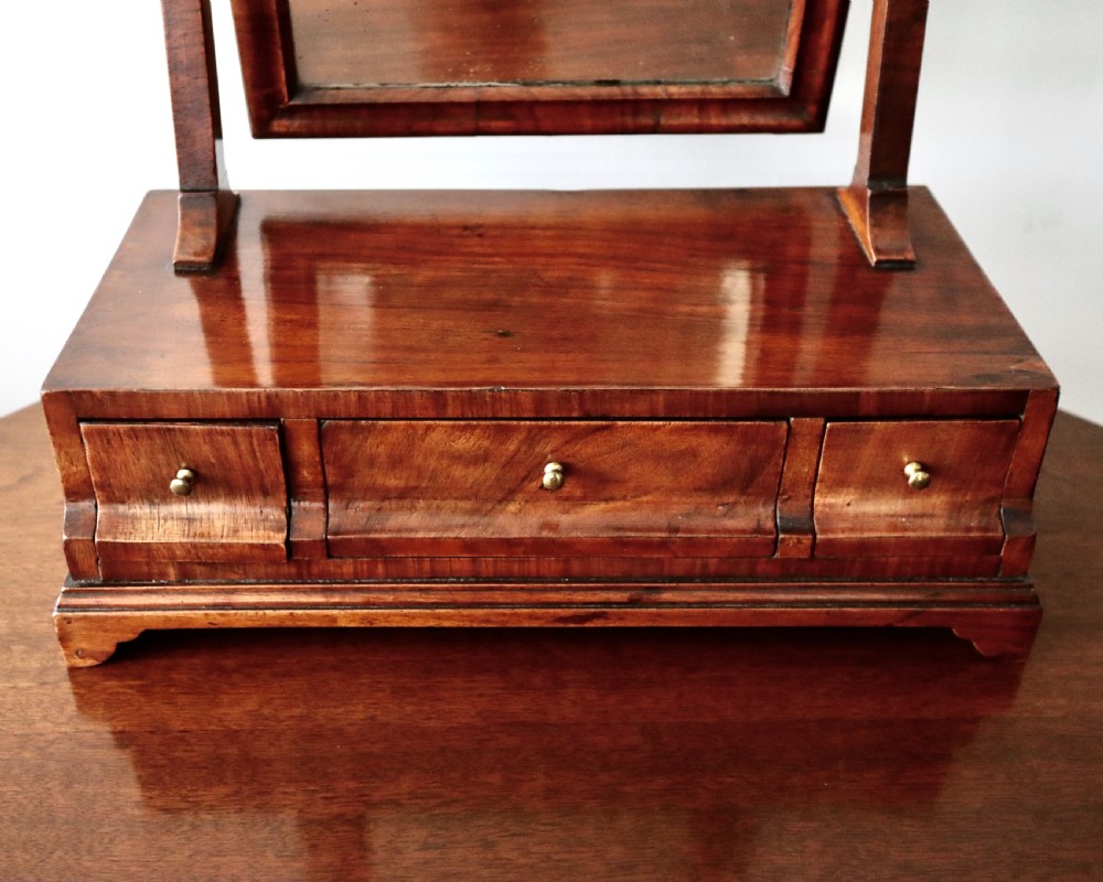 A walnut tabletop toilet mirror featuring an arched swing frame with turned finials mounted above a plinth base containing three small drawers with brass knobs. View III