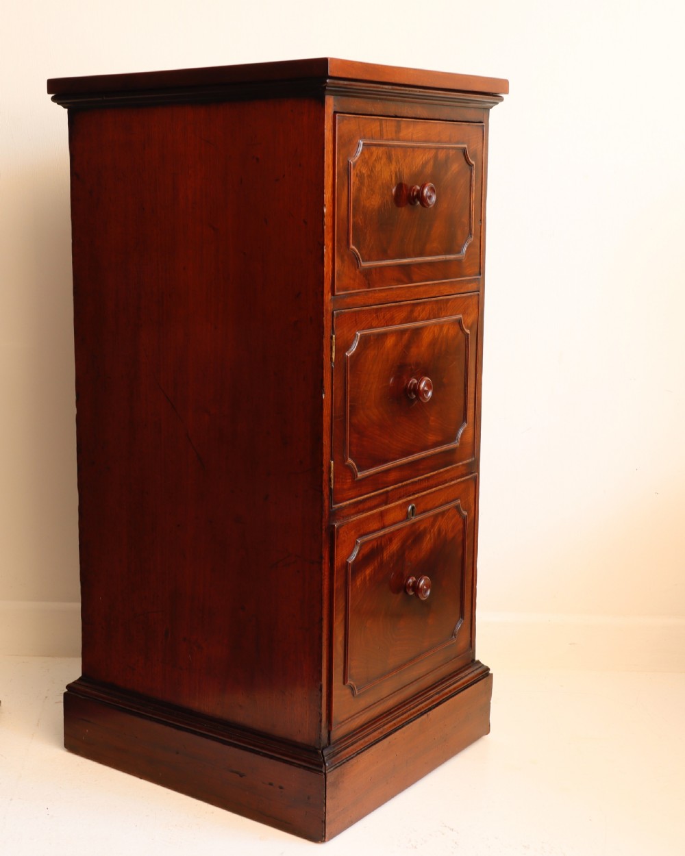 A tall antique flame mahogany cellarette cabinet styled as a chest with three faux drawer fronts, turned wooden knobs, and a solid plinth base. View VII