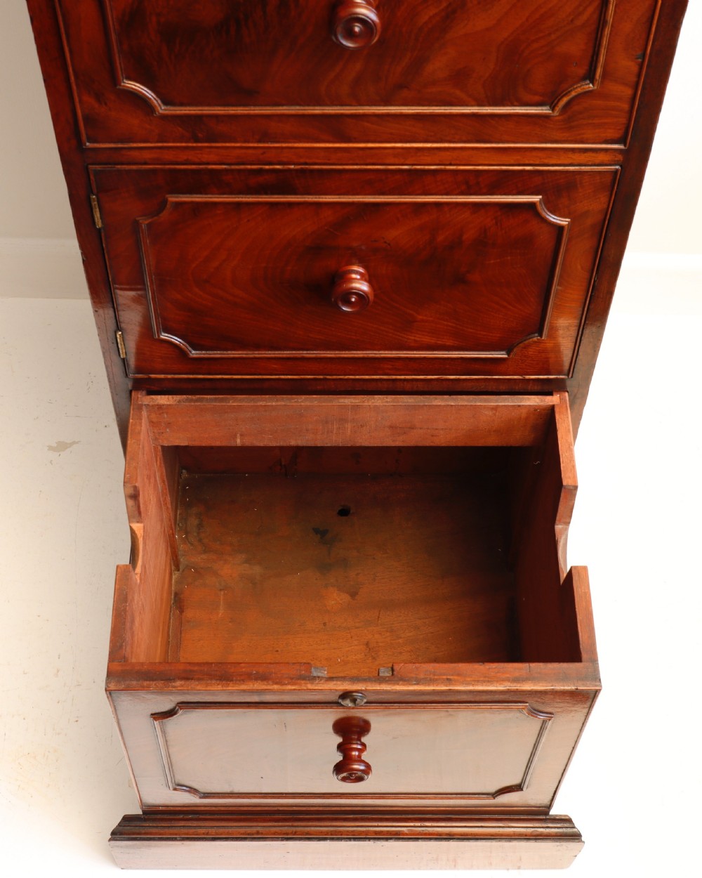 A tall antique flame mahogany cellarette cabinet styled as a chest with three faux drawer fronts, turned wooden knobs, and a solid plinth base. View X