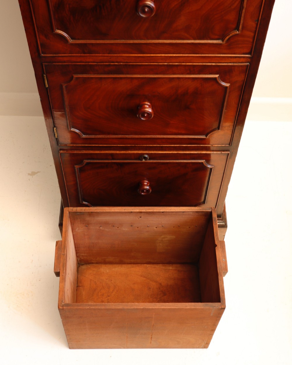 A tall antique flame mahogany cellarette cabinet styled as a chest with three faux drawer fronts, turned wooden knobs, and a solid plinth base. View XI
