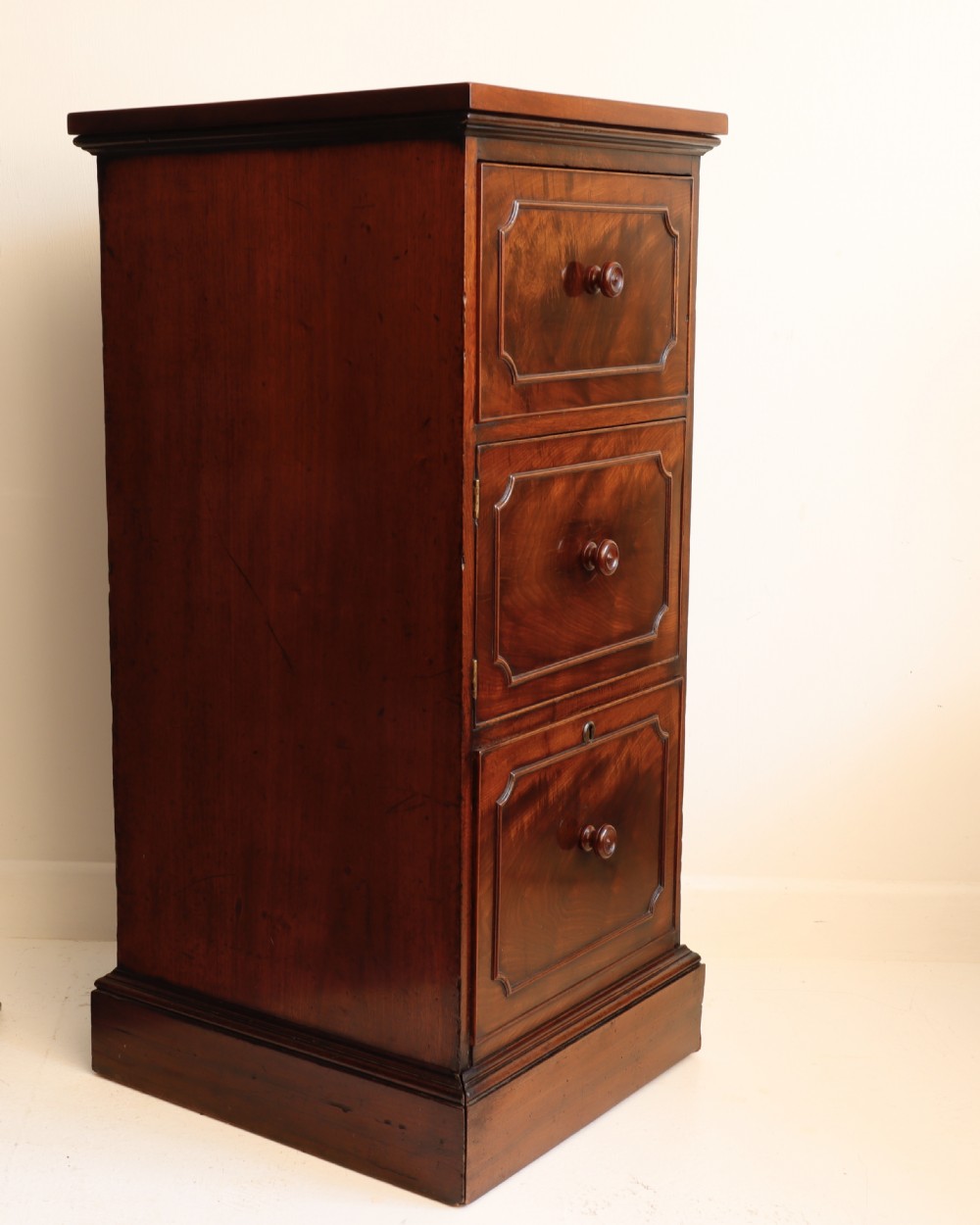 A tall antique flame mahogany cellarette cabinet styled as a chest with three faux drawer fronts, turned wooden knobs, and a solid plinth base.
