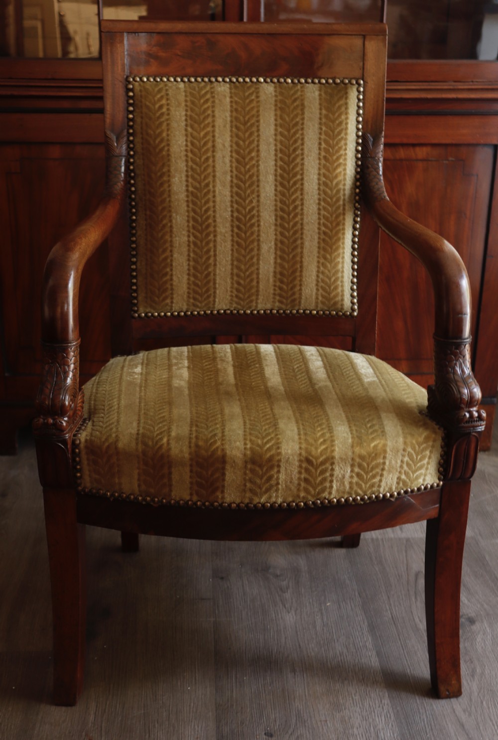 An antique carved mahogany armchair featuring striped velvet upholstery, swept arms with detailed carvings, and brass stud trim.