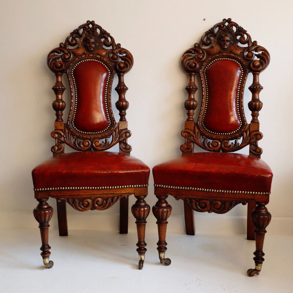 A pair of Victorian carved oak side chairs featuring red leather studded upholstery, carved cresting, and turned front legs on casters.