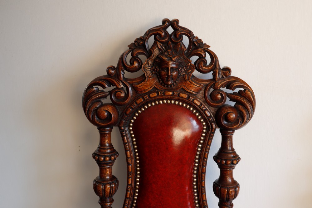 A pair of Victorian carved oak side chairs featuring red leather studded upholstery, carved cresting, and turned front legs on casters. View II