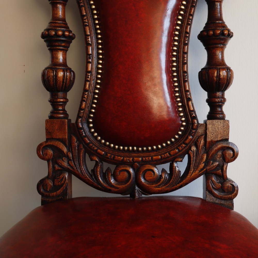 A pair of Victorian carved oak side chairs featuring red leather studded upholstery, carved cresting, and turned front legs on casters. View V