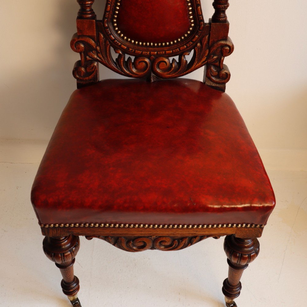 A pair of Victorian carved oak side chairs featuring red leather studded upholstery, carved cresting, and turned front legs on casters. View VII