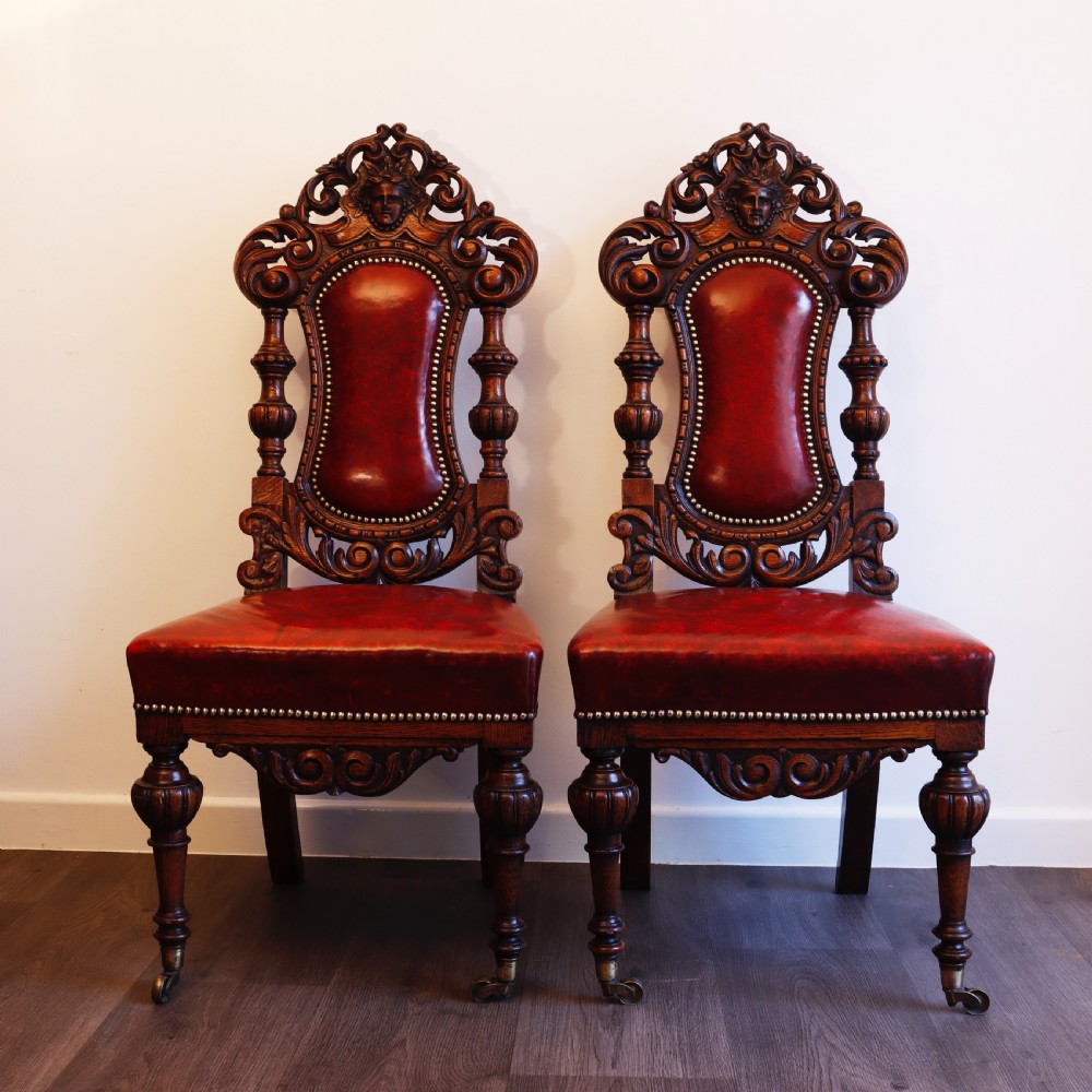 A pair of Victorian carved oak side chairs featuring red leather studded upholstery, carved cresting, and turned front legs on casters. View VIII
