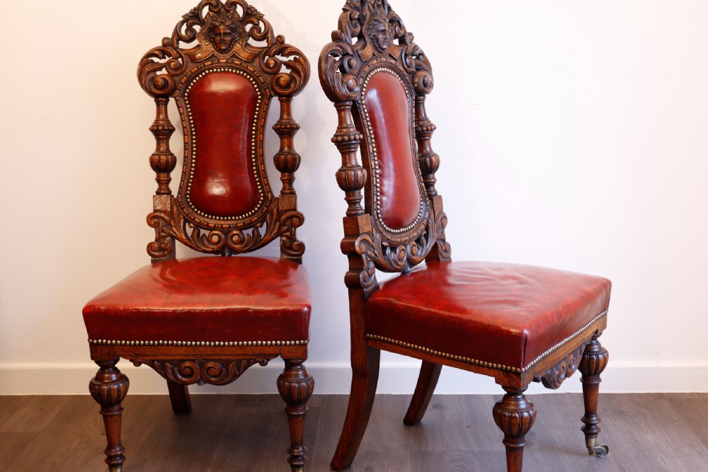A pair of Victorian carved oak side chairs featuring red leather studded upholstery, carved cresting, and turned front legs on casters. View X
