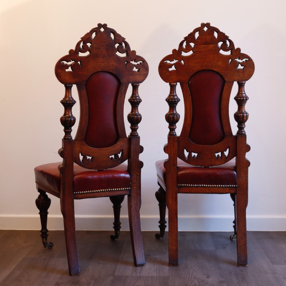 A pair of Victorian carved oak side chairs featuring red leather studded upholstery, carved cresting, and turned front legs on casters. View XI