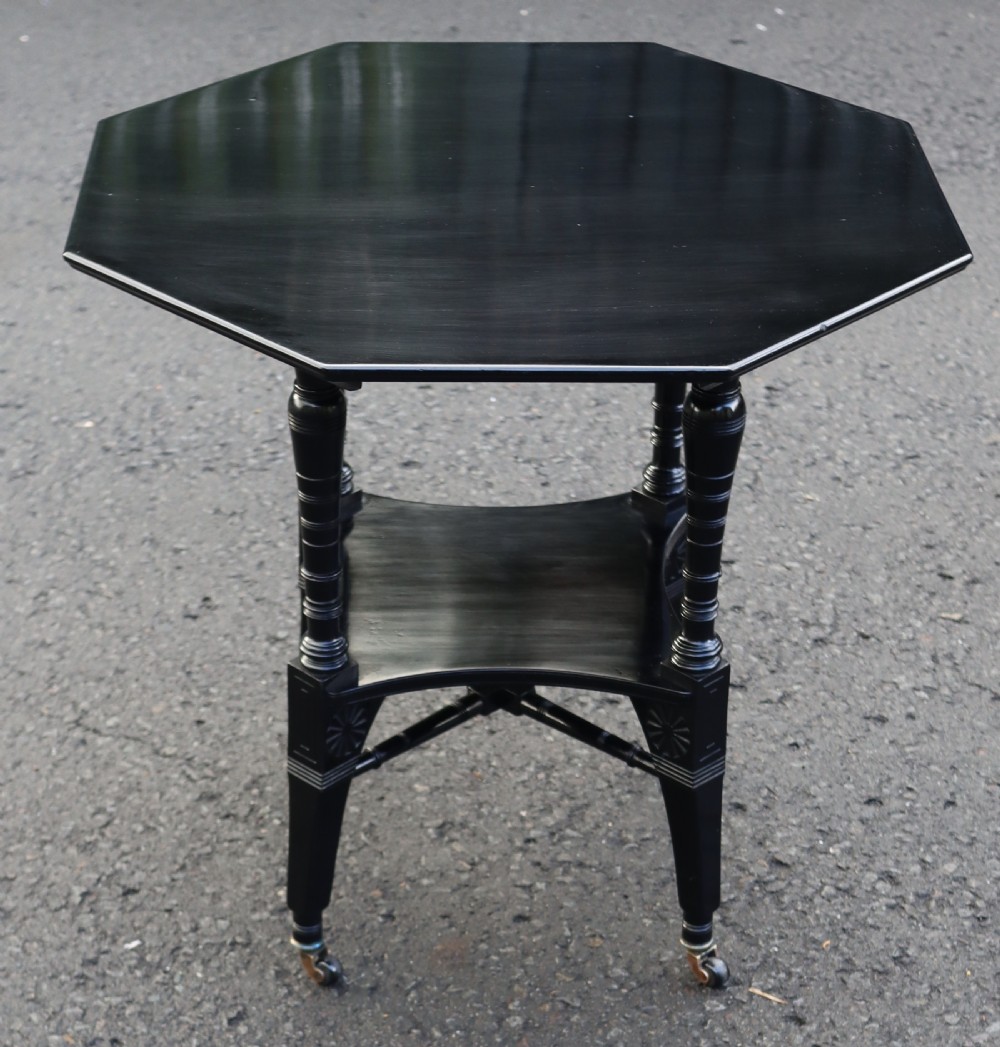 An octagonal ebonised wood two-tier occasional table featuring turned legs, carved relief motifs, cross stretchers, and brass castors.