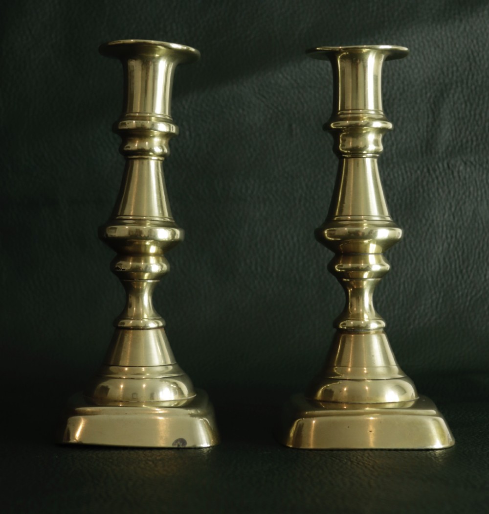 A matching pair of cast brass candlesticks featuring knopped turned columns and square bases resting on a dark surface.