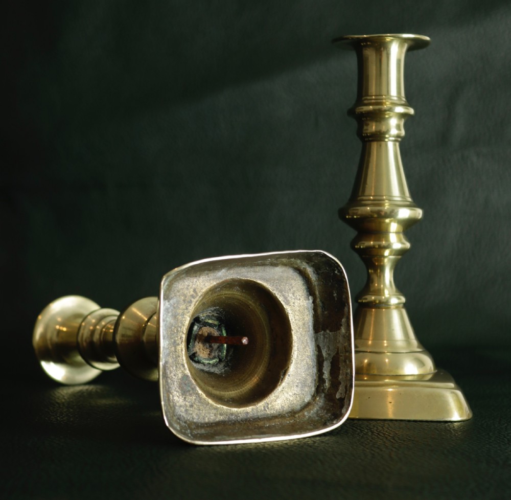 A matching pair of cast brass candlesticks featuring knopped turned columns and square bases resting on a dark surface. View II