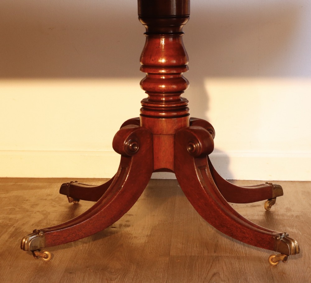 An antique flame mahogany dining table featuring a rectangular crossbanded top with rounded corners supported by a central carved pedestal with four splayed legs terminating in brass castors. View III