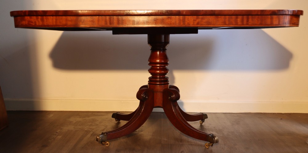 An antique flame mahogany dining table featuring a rectangular crossbanded top with rounded corners supported by a central carved pedestal with four splayed legs terminating in brass castors. View VI
