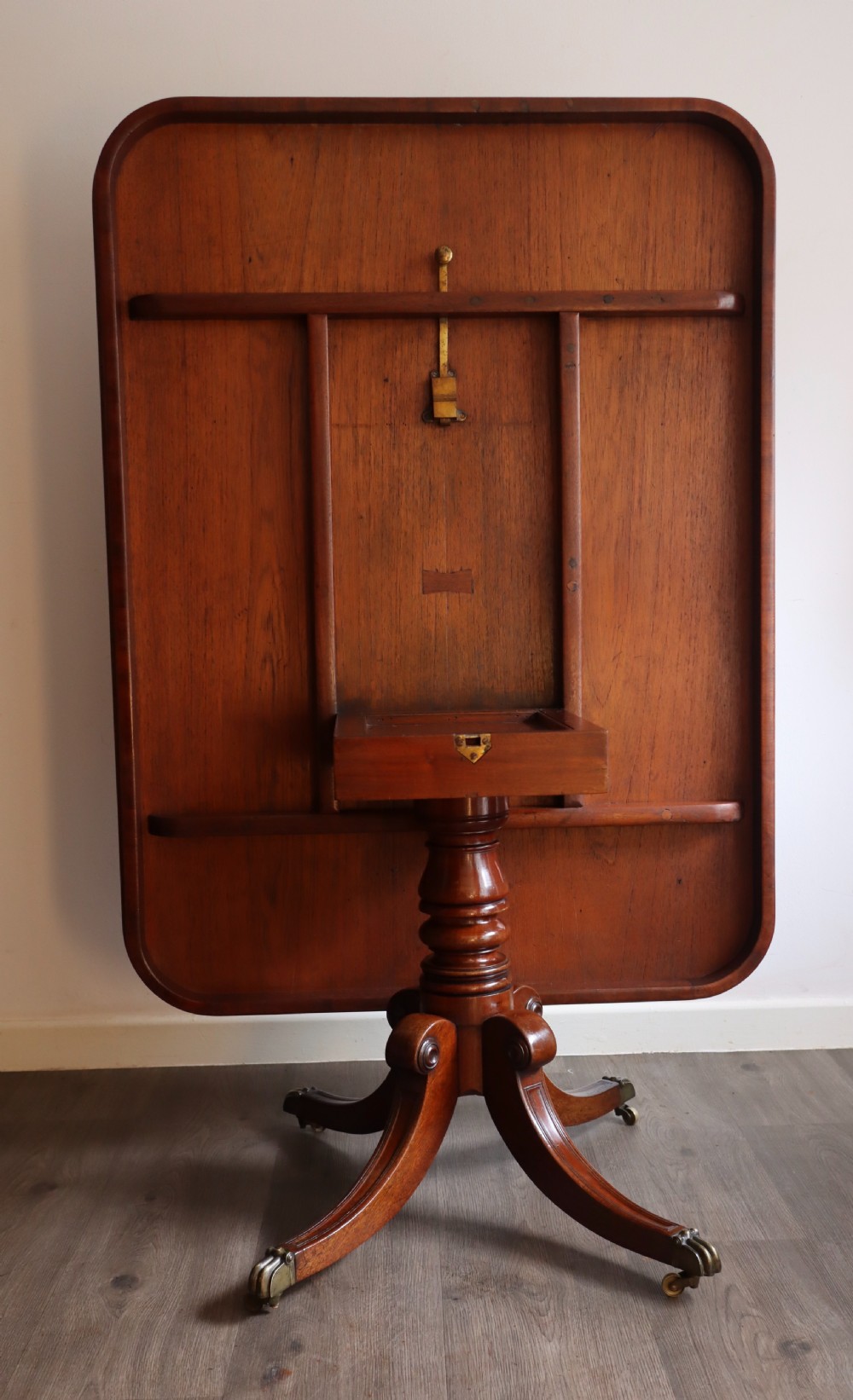 An antique flame mahogany dining table featuring a rectangular crossbanded top with rounded corners supported by a central carved pedestal with four splayed legs terminating in brass castors. View VII