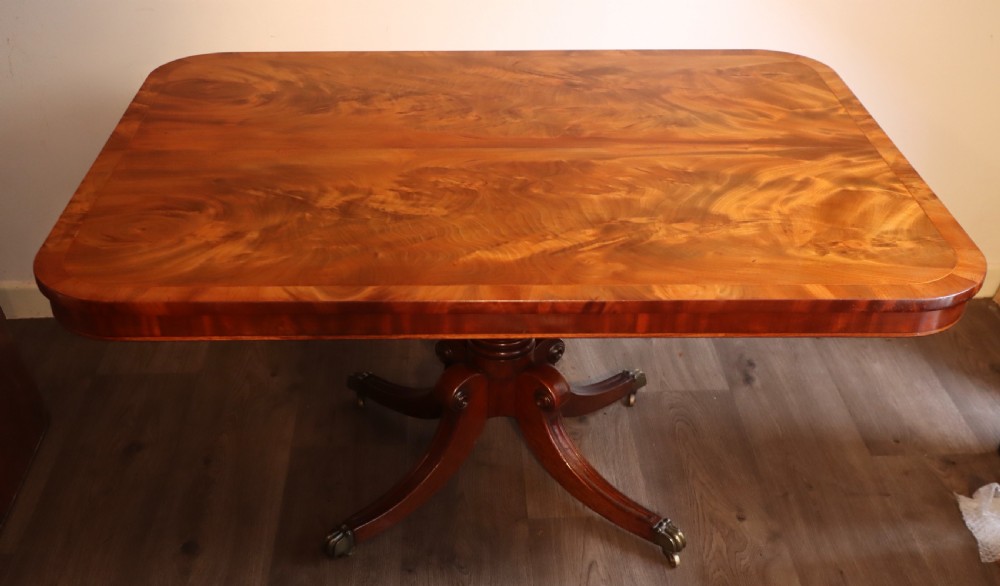 An antique flame mahogany dining table featuring a rectangular crossbanded top with rounded corners supported by a central carved pedestal with four splayed legs terminating in brass castors. View XI