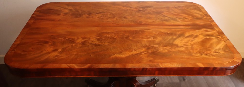 An antique flame mahogany dining table featuring a rectangular crossbanded top with rounded corners supported by a central carved pedestal with four splayed legs terminating in brass castors. View XII