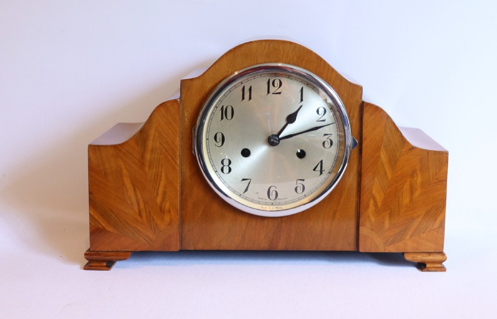 An Art Deco wooden mantel clock featuring an arched case with book-matched veneer and a round silvered dial with Arabic numerals.