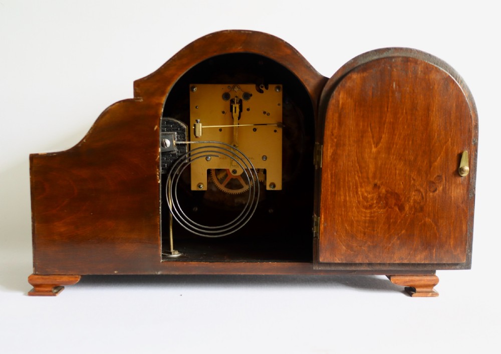An Art Deco wooden mantel clock featuring an arched case with book-matched veneer and a round silvered dial with Arabic numerals. View VII