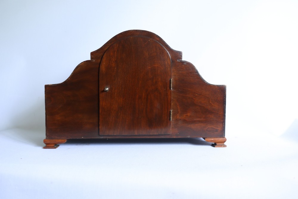 An Art Deco wooden mantel clock featuring an arched case with book-matched veneer and a round silvered dial with Arabic numerals. View X