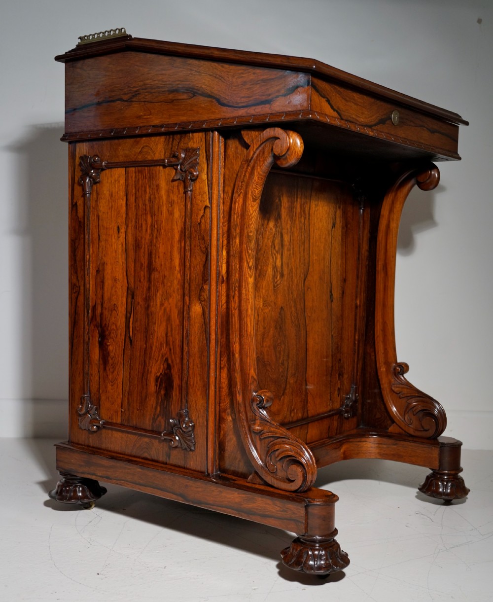 A dark brown rosewood Davenport desk featuring carved scroll front supports, side drawer panelling, and a slanted writing top.