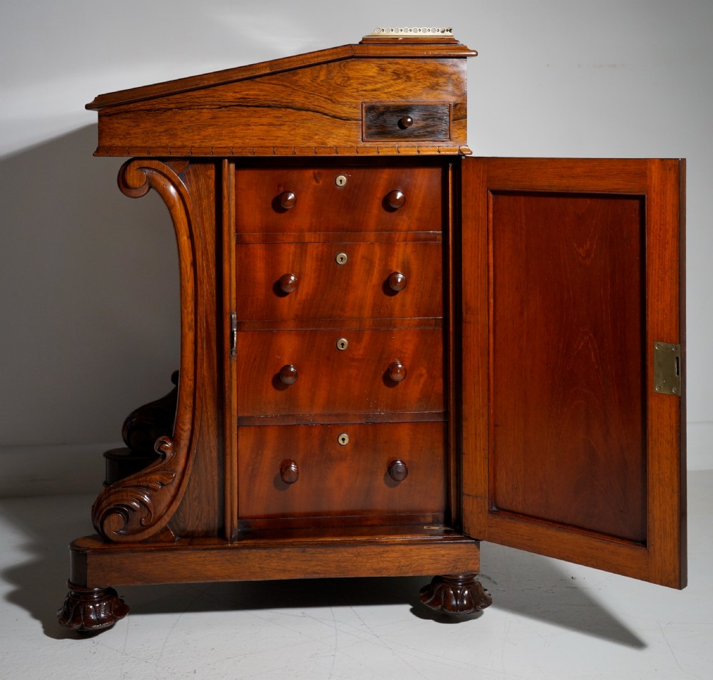 A dark brown rosewood Davenport desk featuring carved scroll front supports, side drawer panelling, and a slanted writing top. View VIII