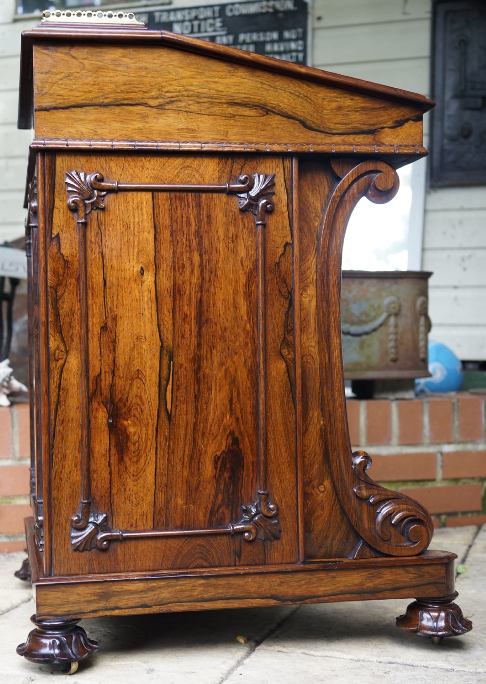 A dark brown rosewood Davenport desk featuring carved scroll front supports, side drawer panelling, and a slanted writing top. View IX