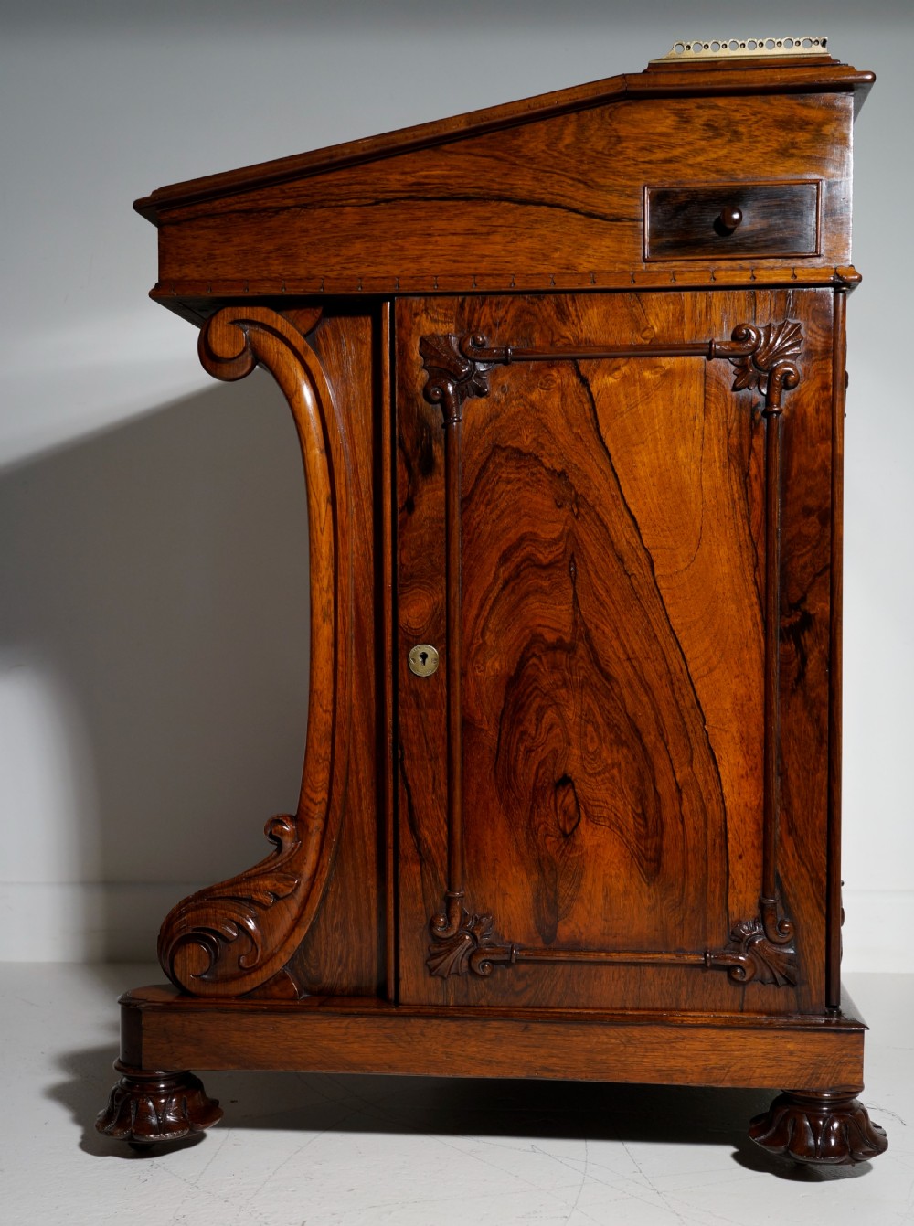 A dark brown rosewood Davenport desk featuring carved scroll front supports, side drawer panelling, and a slanted writing top. View XI
