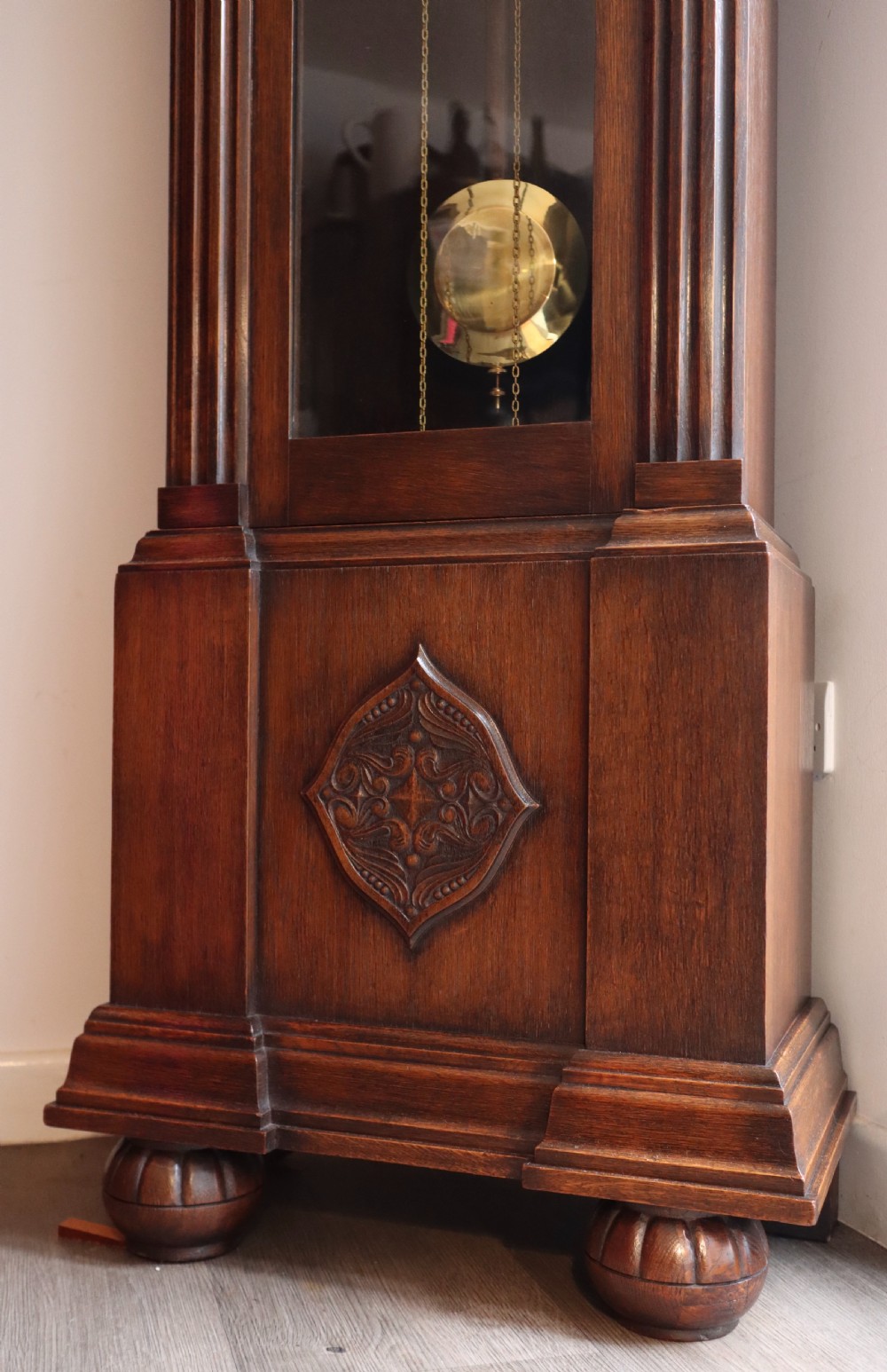 A tall wooden longcase clock featuring fluted corner columns, brass pendulum and weights visible through a glass door, and a circular clock dial with Arabic numerals. View VI