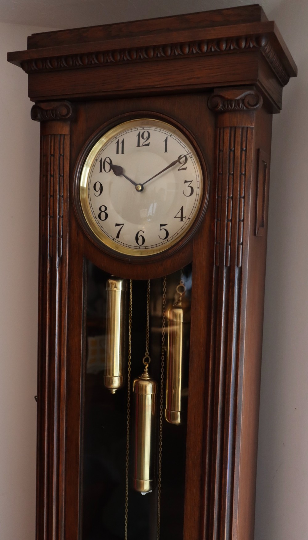 A tall wooden longcase clock featuring fluted corner columns, brass pendulum and weights visible through a glass door, and a circular clock dial with Arabic numerals. View VII