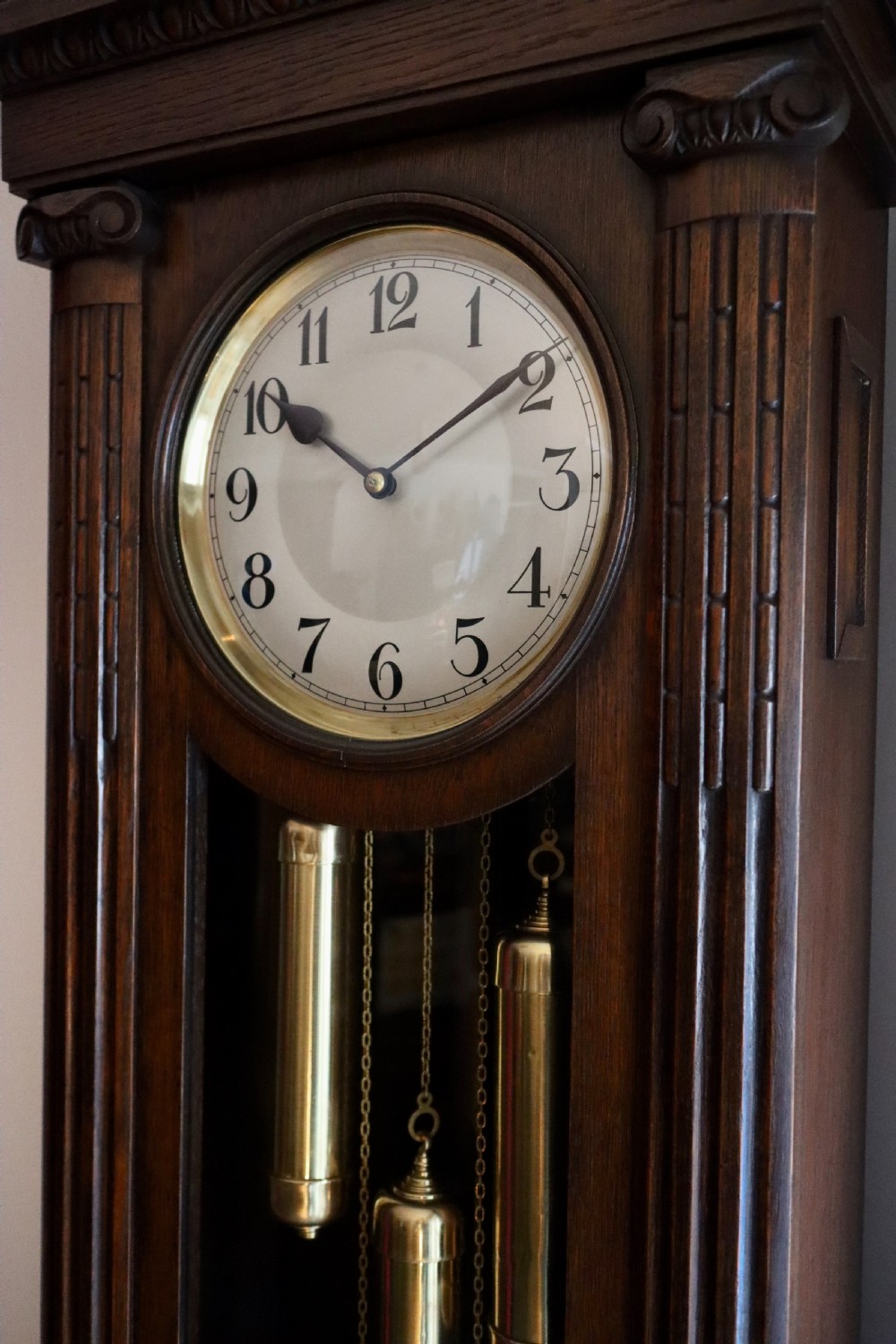A tall wooden longcase clock featuring fluted corner columns, brass pendulum and weights visible through a glass door, and a circular clock dial with Arabic numerals. View VIII