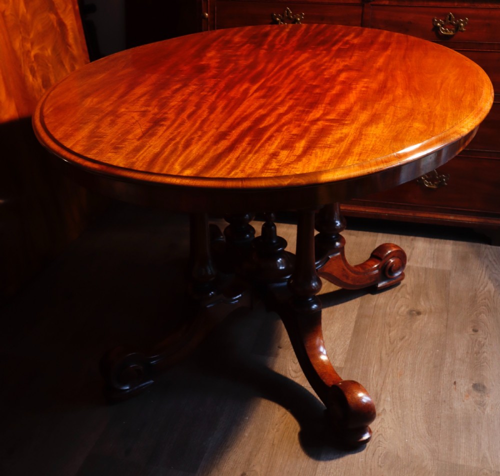 A circular Victorian flame mahogany dining table resting on a turned pedestal with four carved scroll legs.