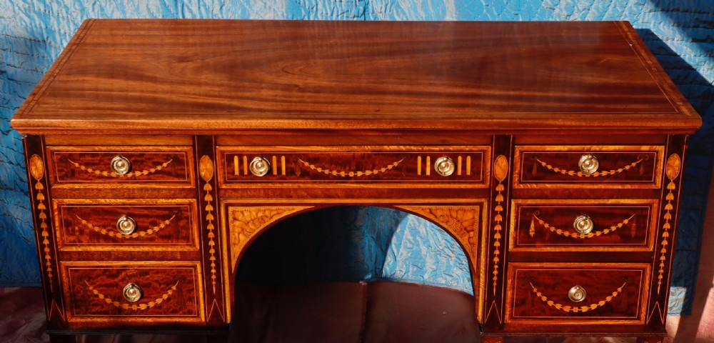 A mahogany writing desk featuring floral marquetry swags, seven drawers with brass handles, a central arch kneehole, and tapered legs on brass castors. View V
