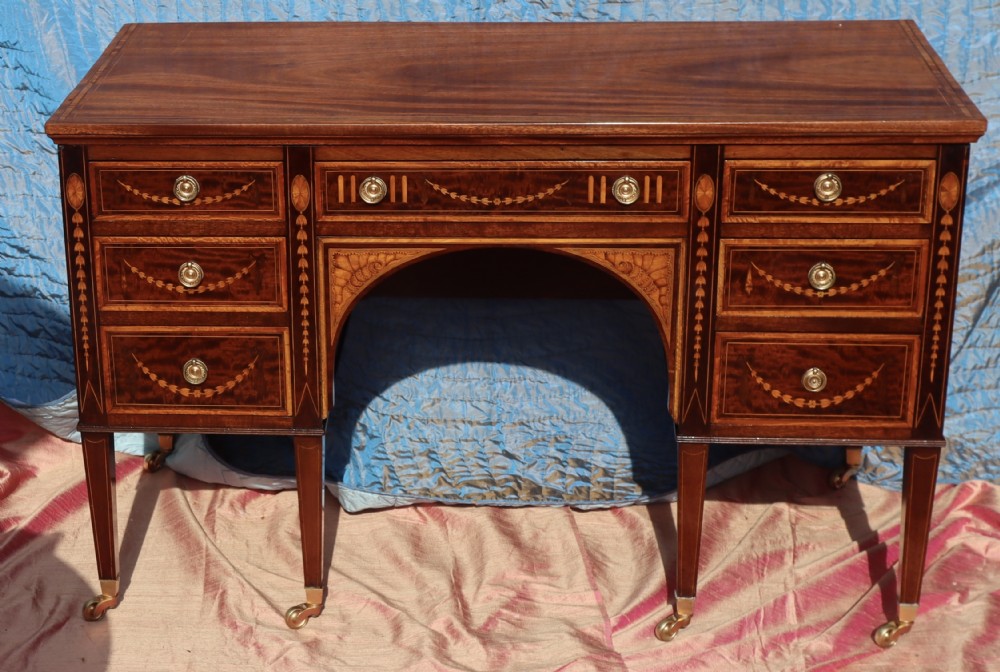 A mahogany writing desk featuring floral marquetry swags, seven drawers with brass handles, a central arch kneehole, and tapered legs on brass castors.