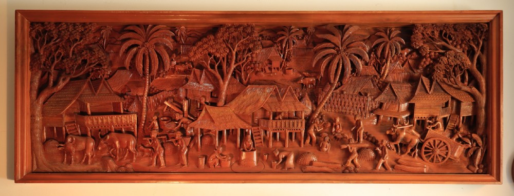 A large rectangular hand-carved teak wall panel depicting a detailed rural village scene with stilt houses, figures, bullocks, and palm trees.