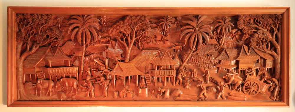 A large rectangular hand-carved teak wall panel depicting a detailed rural village scene with stilt houses, figures, bullocks, and palm trees. View II