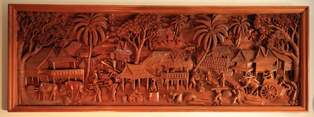 A large rectangular hand-carved teak wall panel depicting a detailed rural village scene with stilt houses, figures, bullocks, and palm trees. View III
