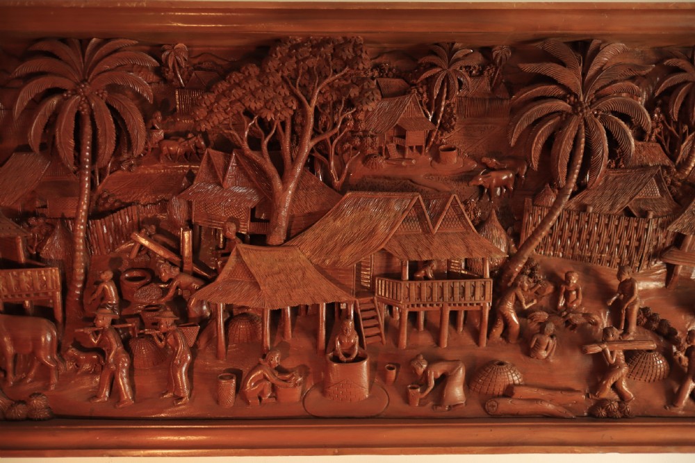 A large rectangular hand-carved teak wall panel depicting a detailed rural village scene with stilt houses, figures, bullocks, and palm trees. View V