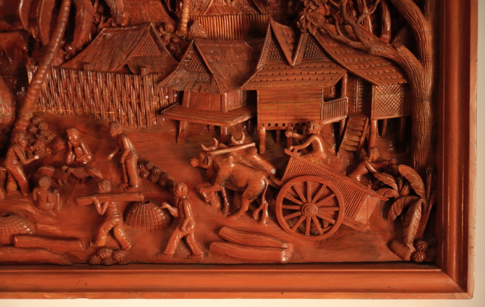 A large rectangular hand-carved teak wall panel depicting a detailed rural village scene with stilt houses, figures, bullocks, and palm trees. View VI