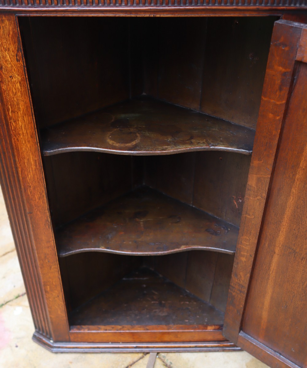 An antique oak corner cupboard featuring a single door with central oval starburst inlay, fluted pilasters, brass H-hinges, and a dentil moulding cornice. View VI